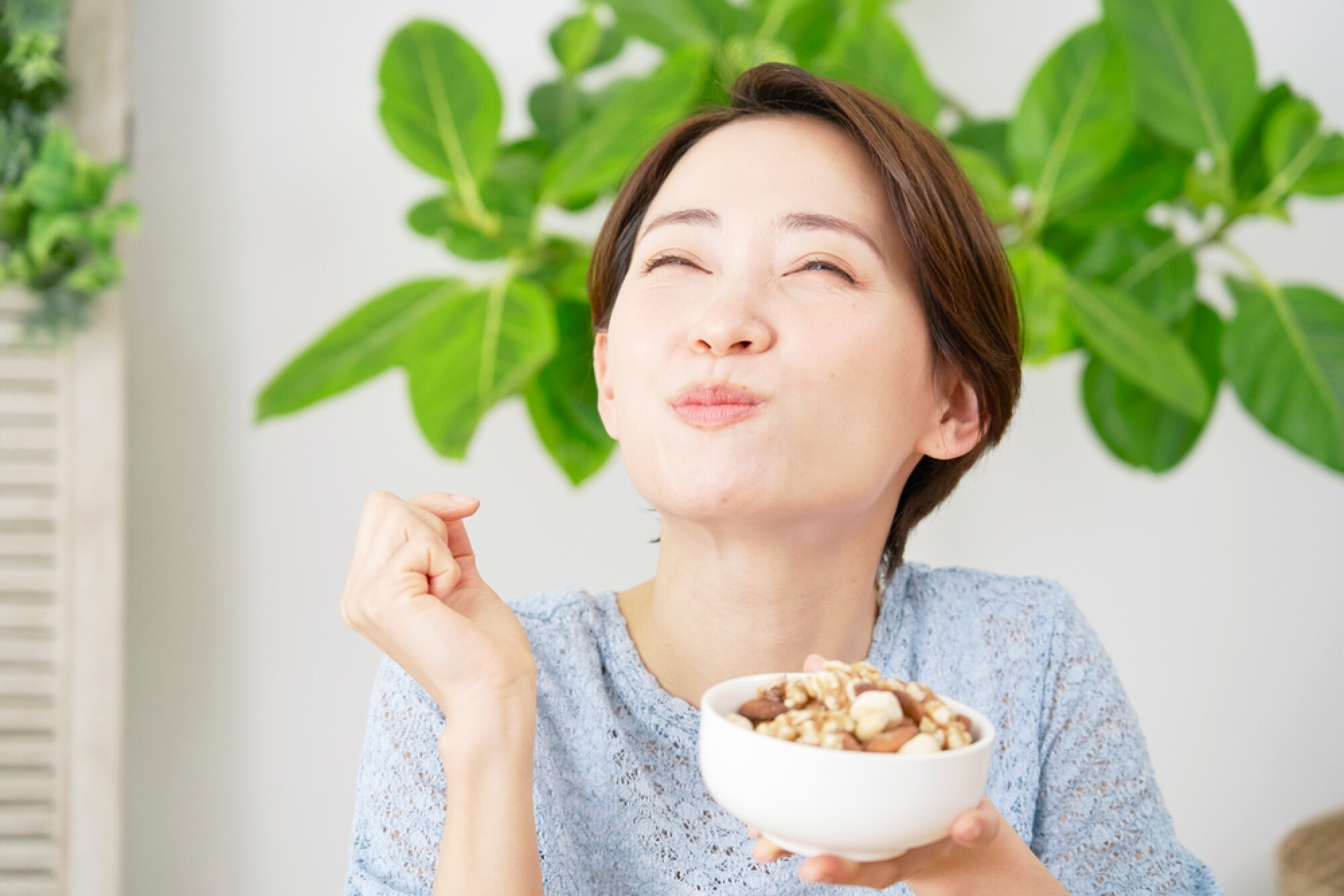 一位女性正在品嚐一碗健康的堅果燕麥，面帶微笑，享受食物的美味，背景是綠意盎然的室內植物。
