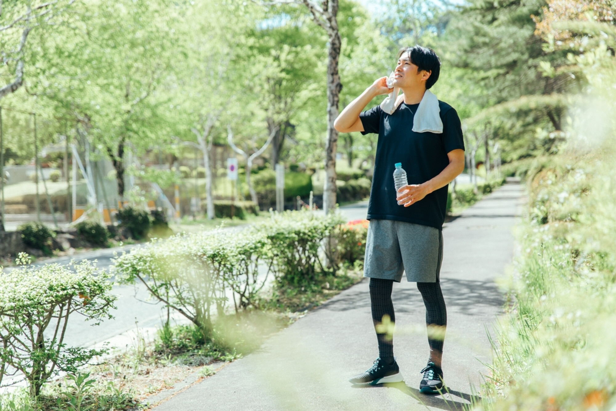  一位穿著運動服的年輕男士在公園的小徑上休息，他一手拿著水瓶，另一手用毛巾擦汗。