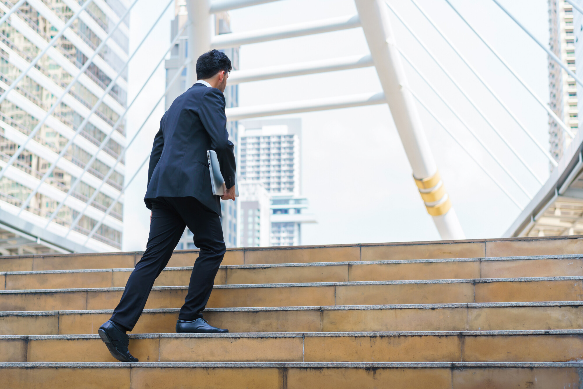 一位穿著西裝的男子拿著手提電腦，正快步走上戶外樓梯，背景是現代都市建築。