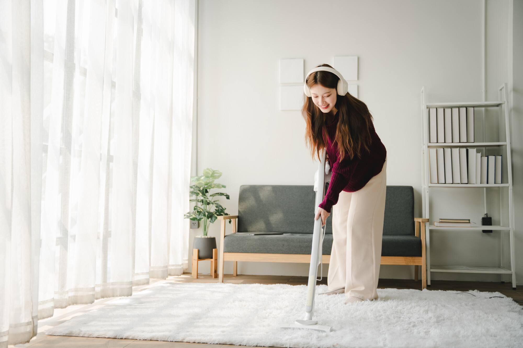 一位女子戴著耳機，穿著紫色毛衣，正在客廳用吸塵器清理地毯，面帶微笑。