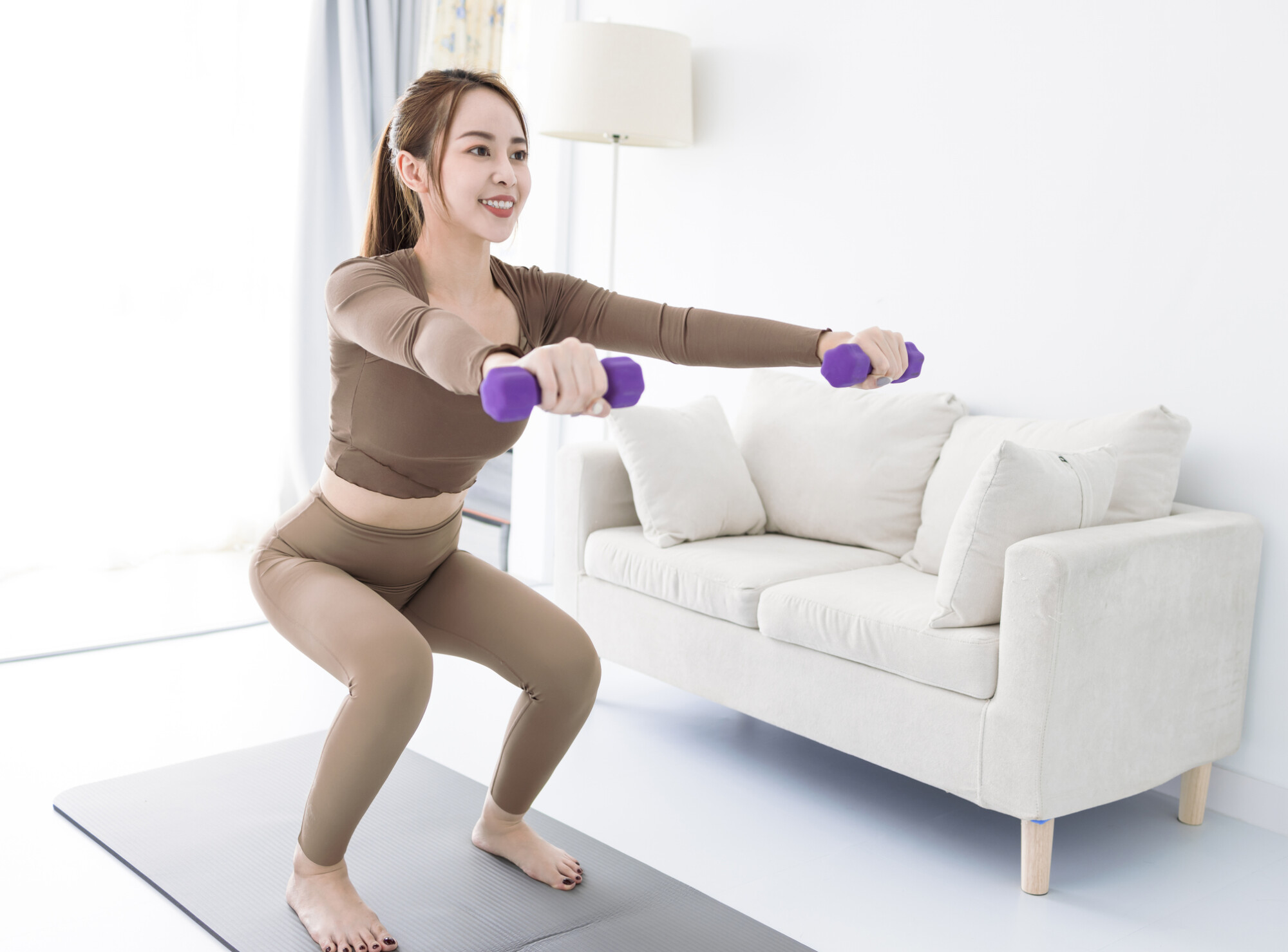 一位面帶微笑的年輕女士正在家中的瑜珈墊上，雙手拿著紫色啞鈴進行深蹲運動。