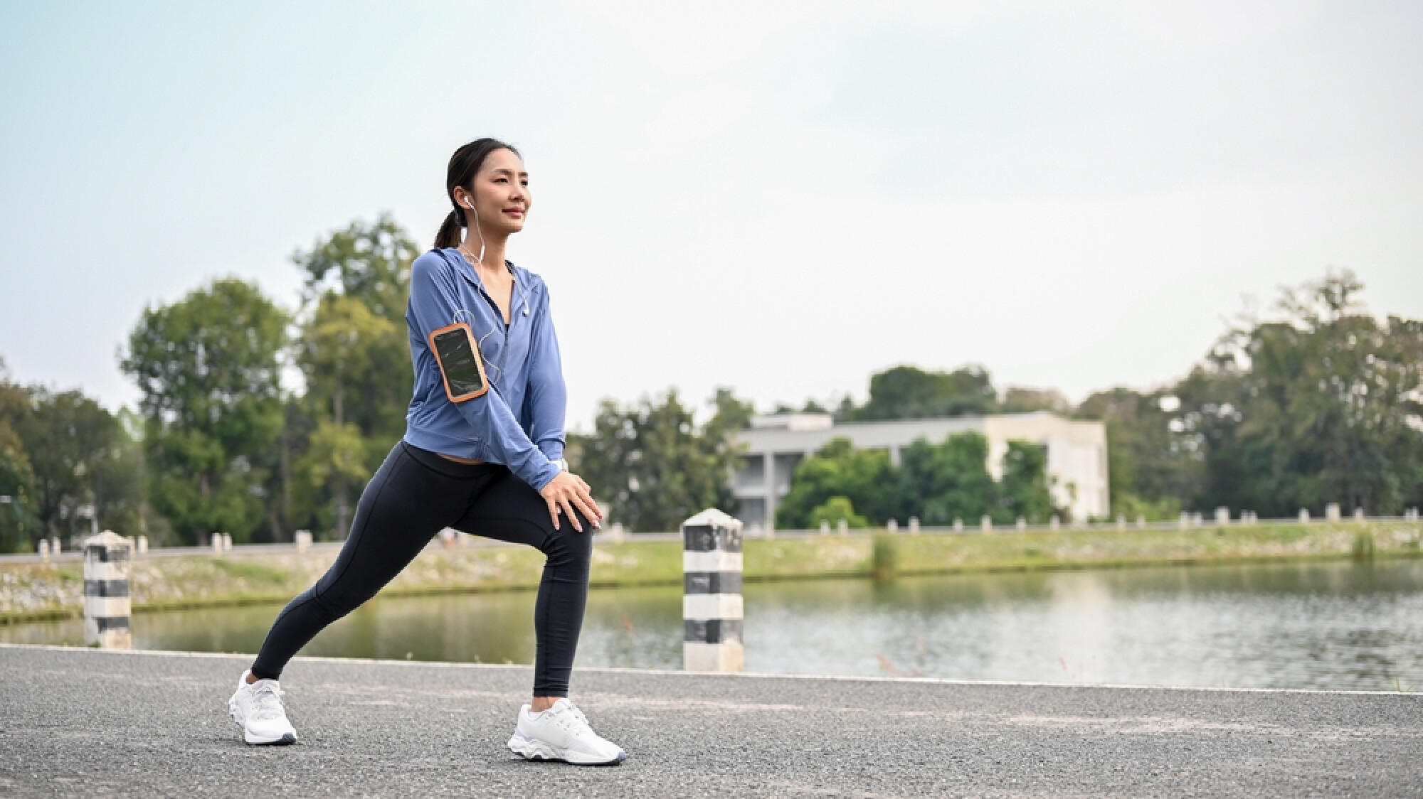 一位穿著藍色運動外套和緊身褲的女士，戴著耳機，在湖邊的小徑上做弓步伸展。