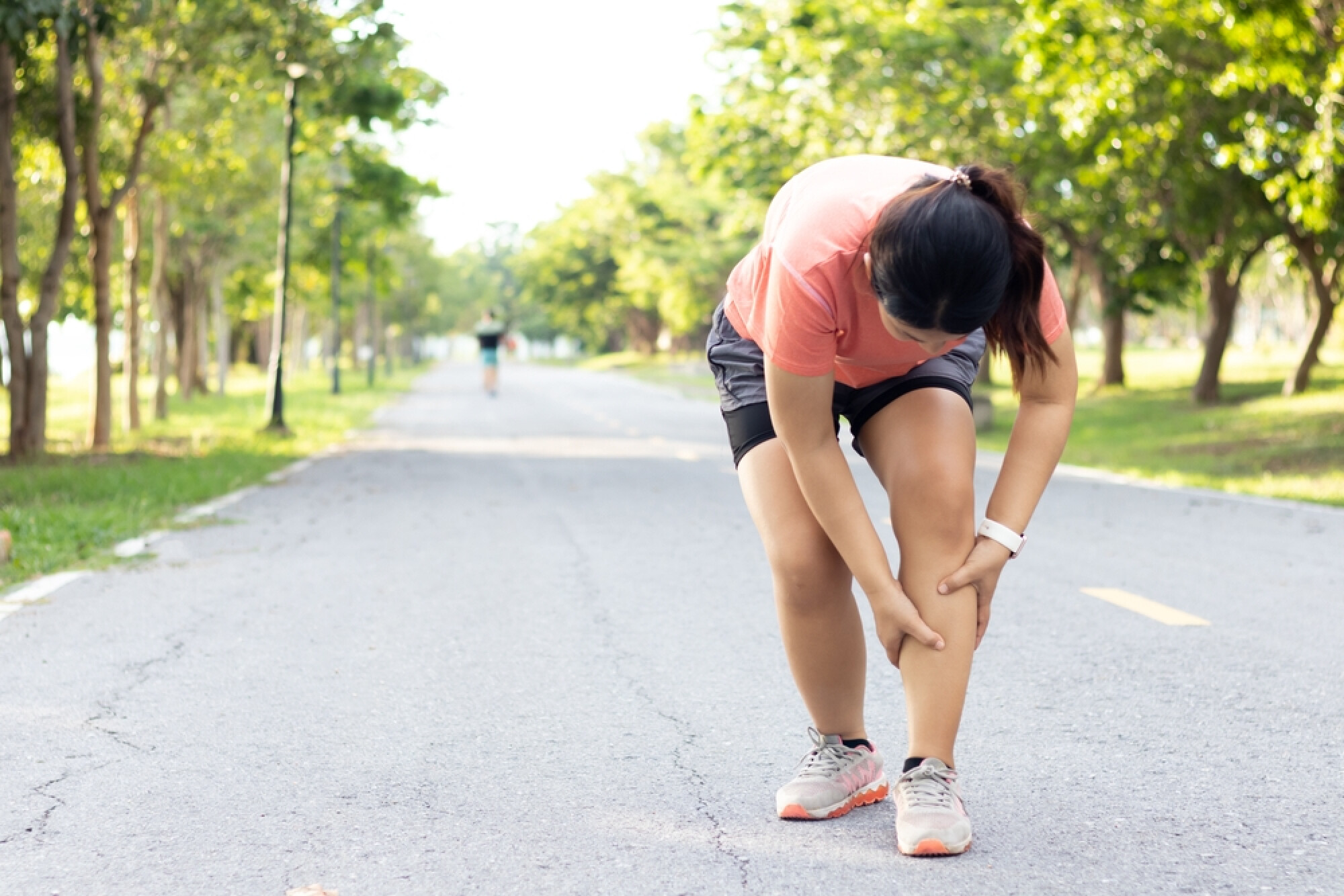 一位女士在綠樹成蔭的公園跑道上停下來，彎下腰痛苦地按摩著她的小腿，似乎是抽筋或受傷了。