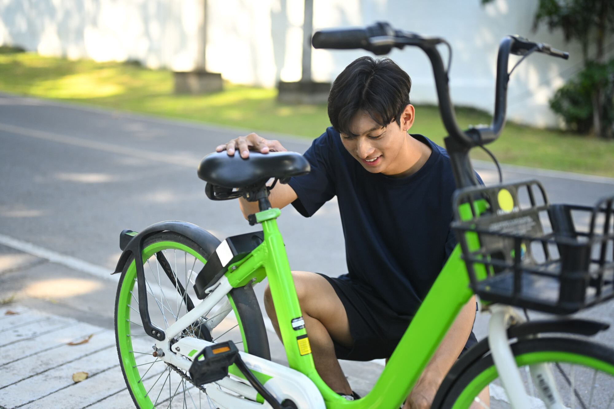 年輕男子蹲在路邊微笑調整綠色共享單車的座椅高度，仔細檢查車輛狀況，為接下來的城市騎行做準備。