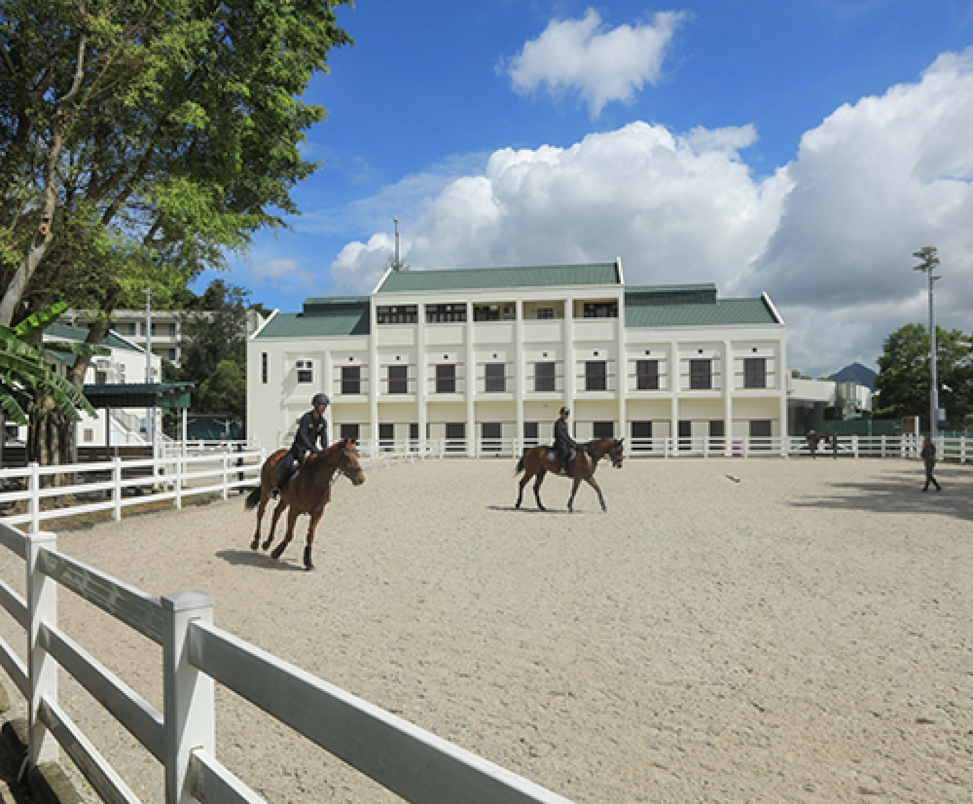 鯉魚門公眾騎術學校的白色主校舍外觀及戶外沙圈，學員正在教練指導下進行馬術訓練。