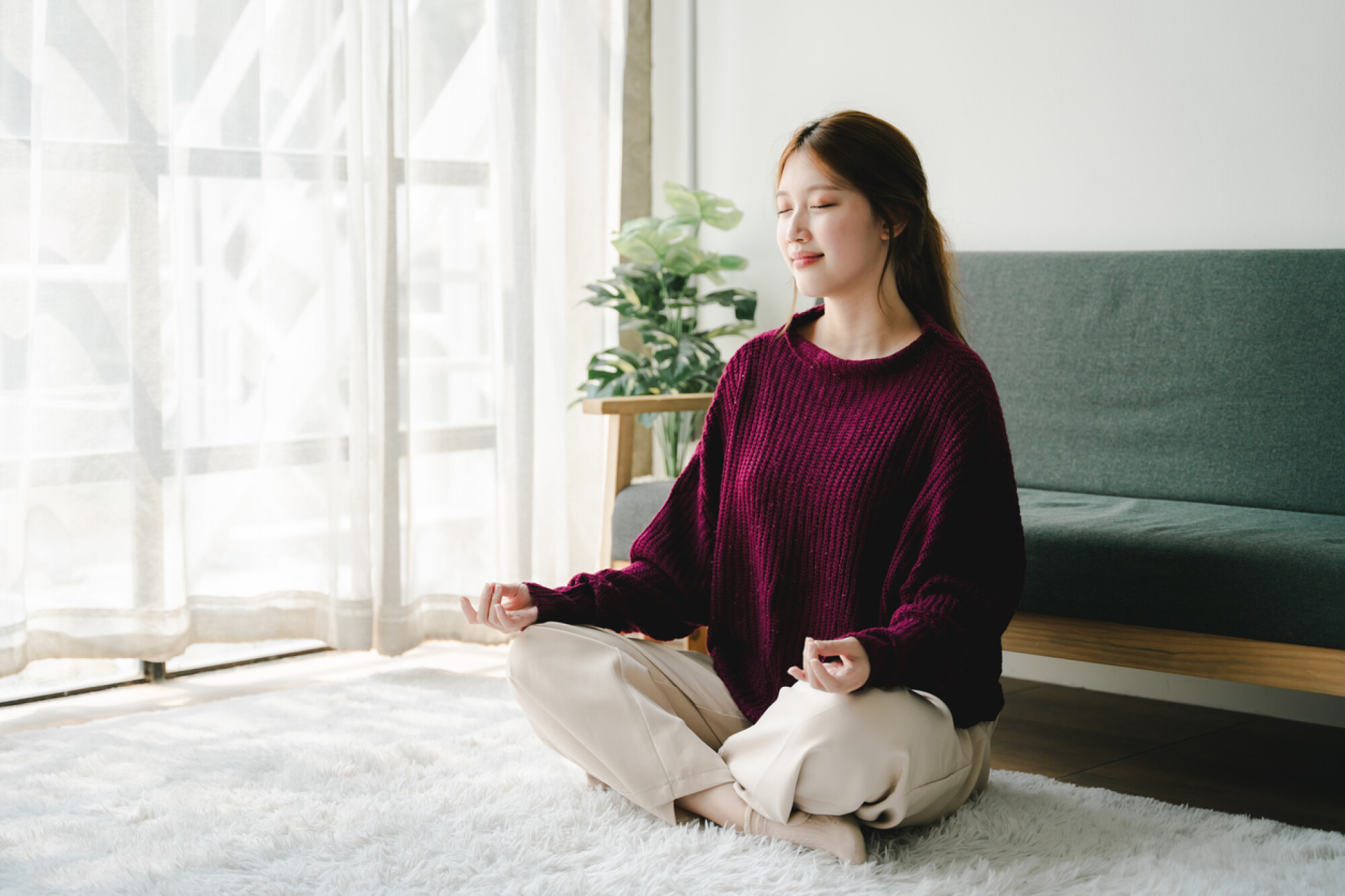 一位穿著酒紅色針織毛衣和米色褲子的女子坐在室內地毯上，雙腿盤坐，雙手放在膝蓋上，眼睛閉著，身旁有綠色植物，陽光從窗戶灑進室內，氣氛溫馨寧靜。