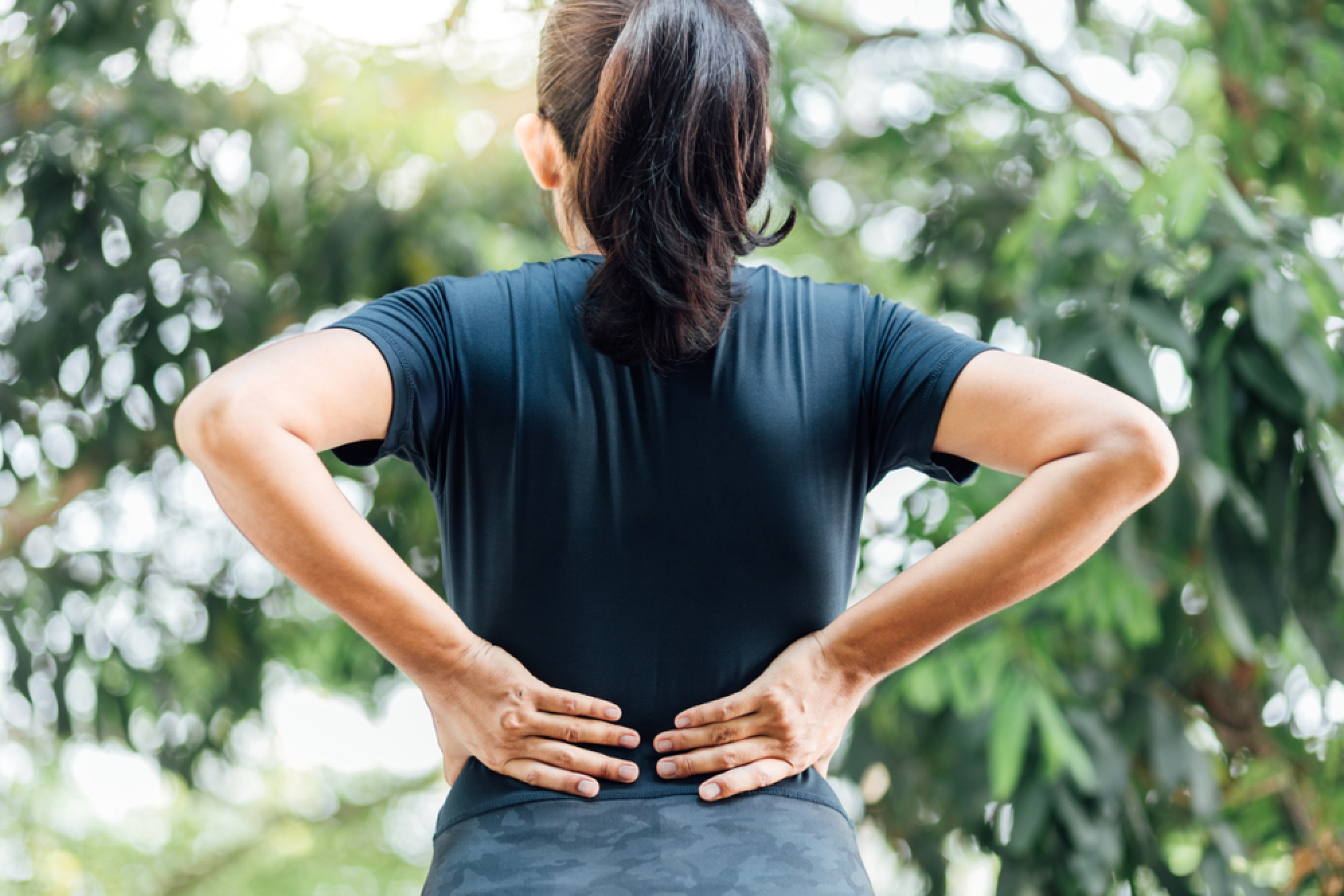 The back of a woman standing outdoors in front of green trees, with her hands on her lower back as if she is experiencing back pain.
