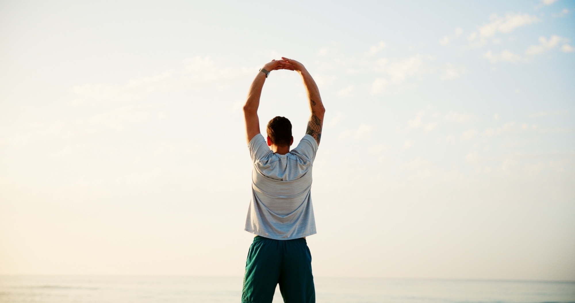 A man with his back to the camera, standing by the sea with his arms stretched overhead, facing a wide sky at sunrise or sunset.