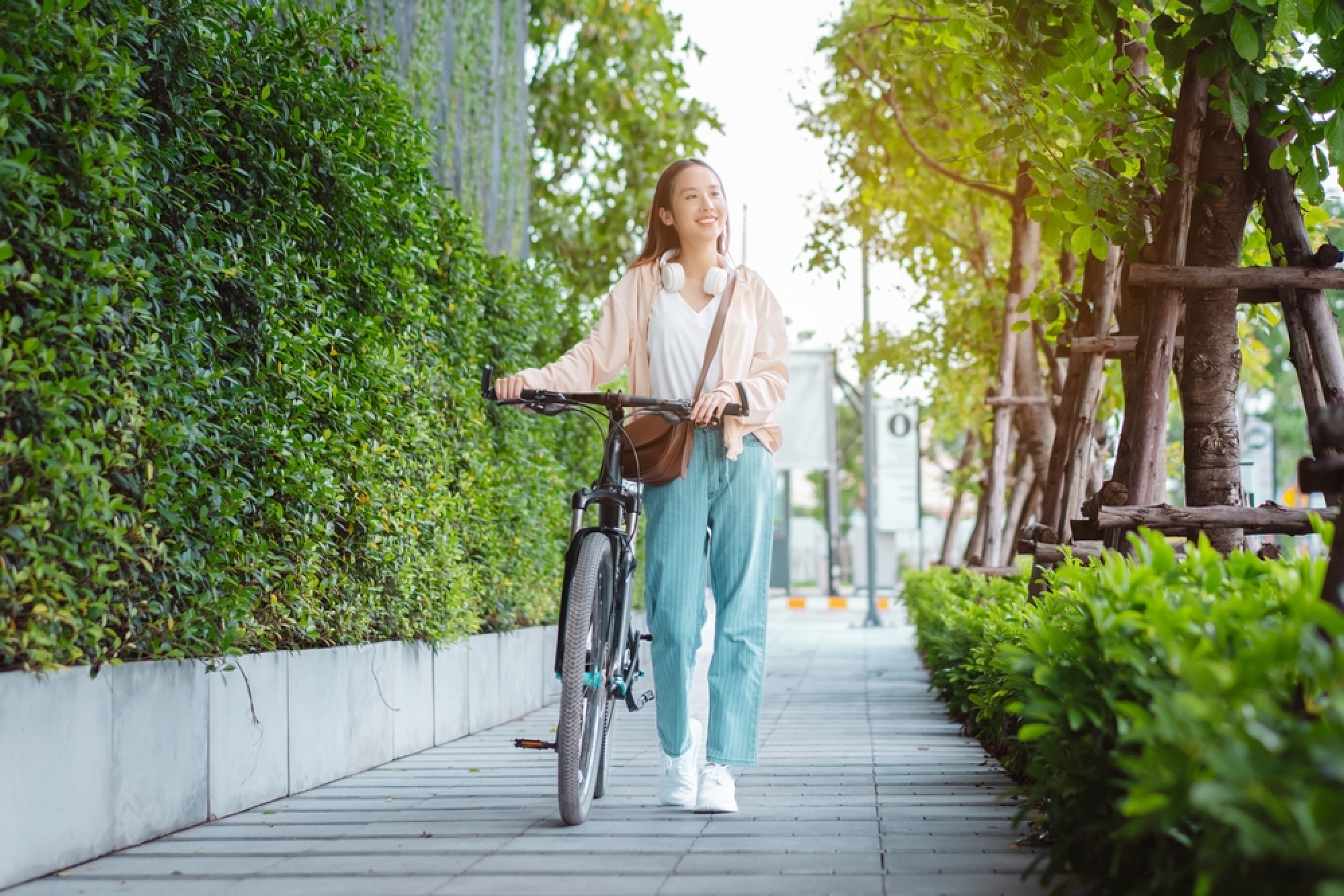 A smiling young woman in casual wear pushes a bicycle along a sidewalk lined with green trees.