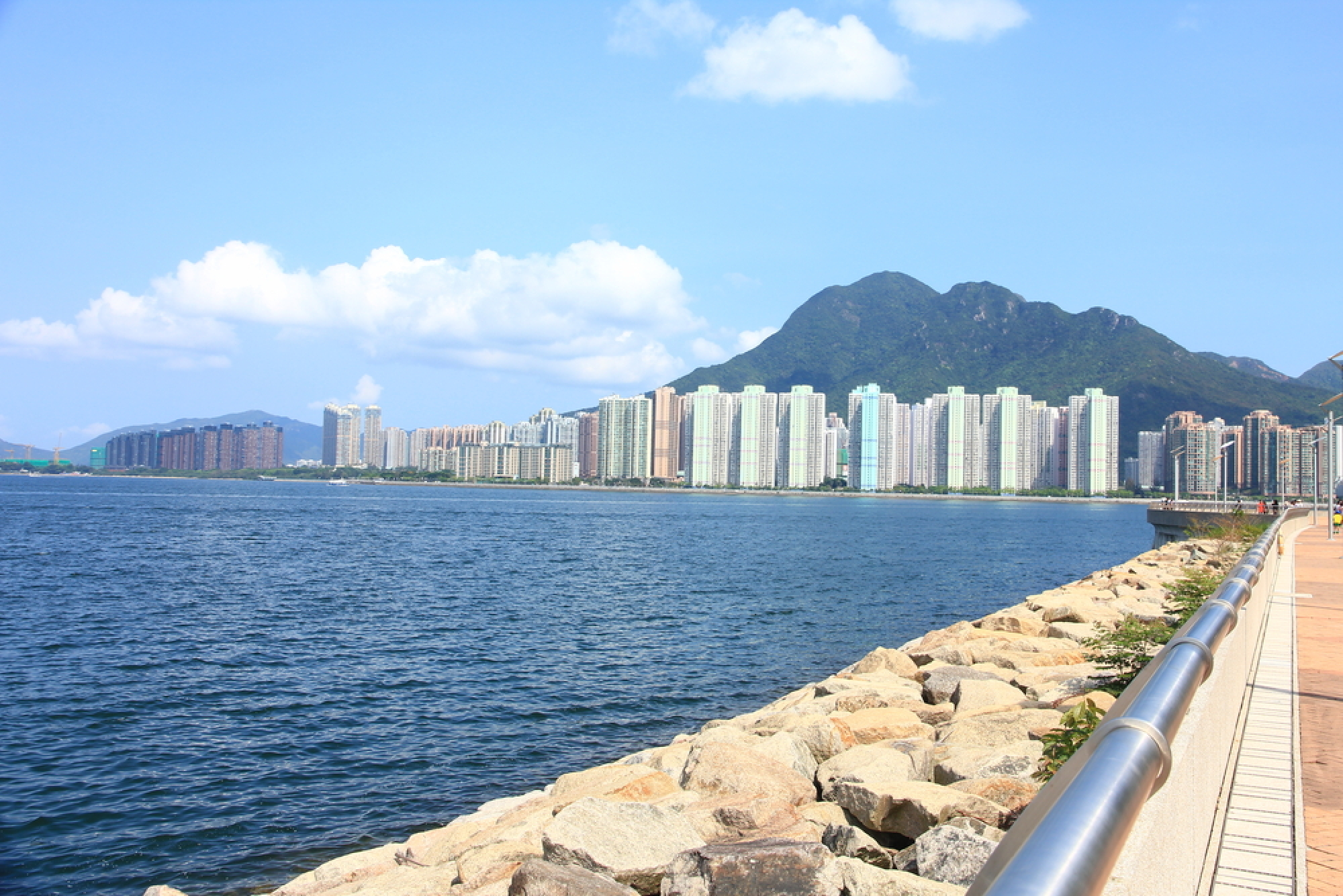 從石砌海濱長廊望向海灣對岸的香港馬鞍山城市景觀，可見沿岸的高樓住宅大廈和後方的翠綠山巒。