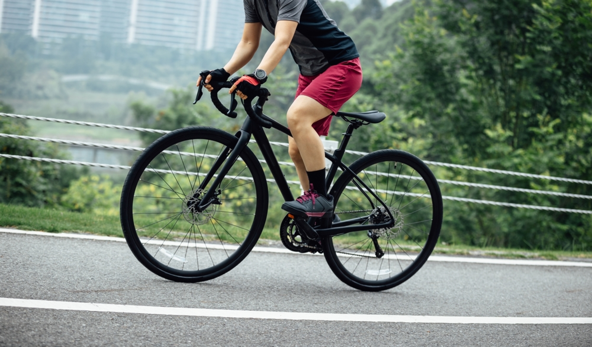 A side close-up of a person riding a black road bike, showing their legs pedaling, with a blurred background of a city and mountains.