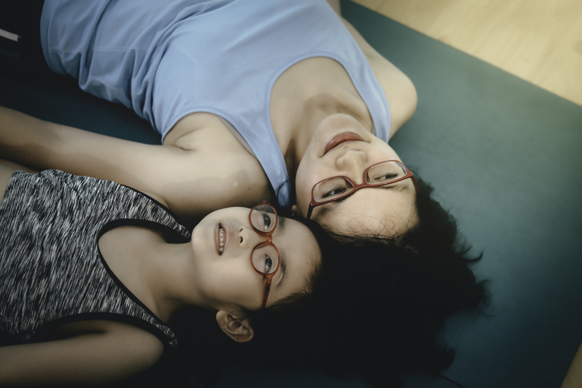 從正上方拍攝，一位戴著眼鏡的母親和女兒並排躺在瑜珈墊上，一起向上看。