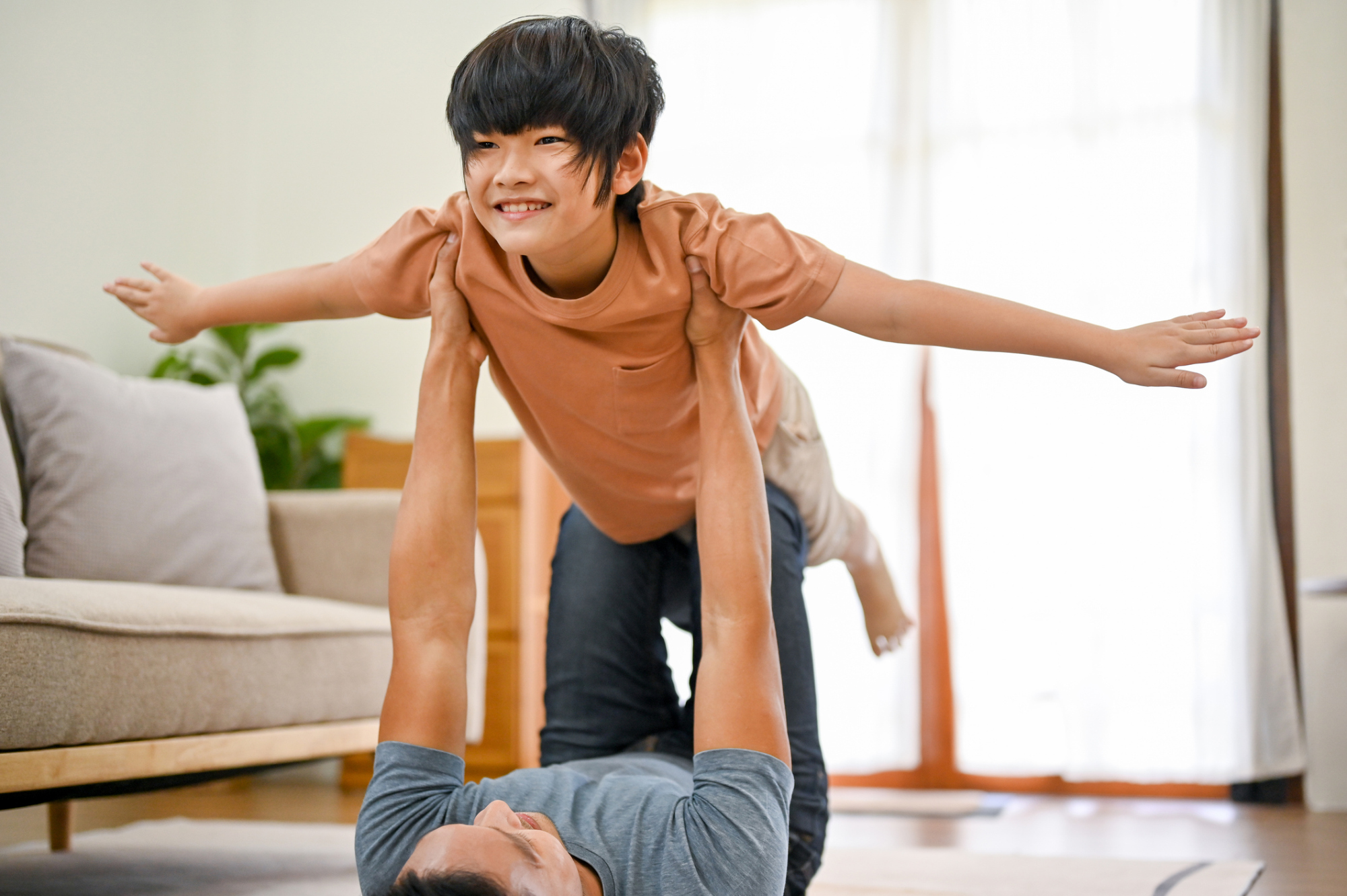 A father lying on the floor at home, lifting his smiling son into the air with his hands, while the son spreads his arms out like an airplane.