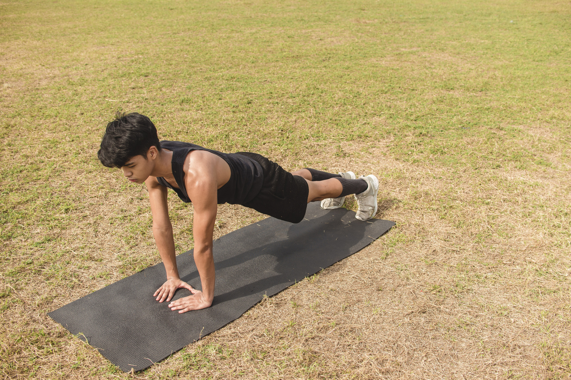A young man on a yoga mat on the grass outdoors, in a high plank position (push-up ready position) with his hands on the ground.