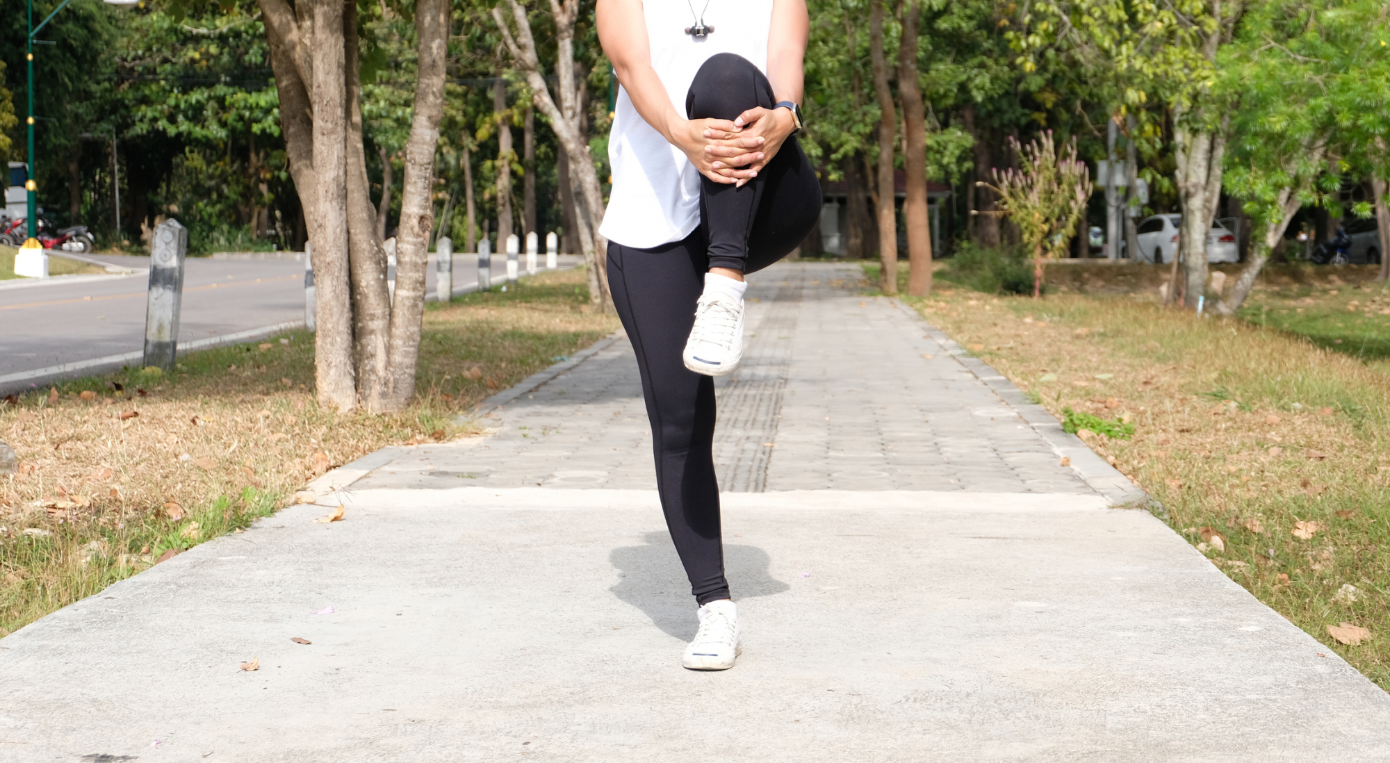 iStock-1580791641.jpg 女子在戶外進行抱膝走 (Knee Hug) 動態熱身,單腿站立並將膝蓋抱向胸口,以伸展臀大肌與髖關節。